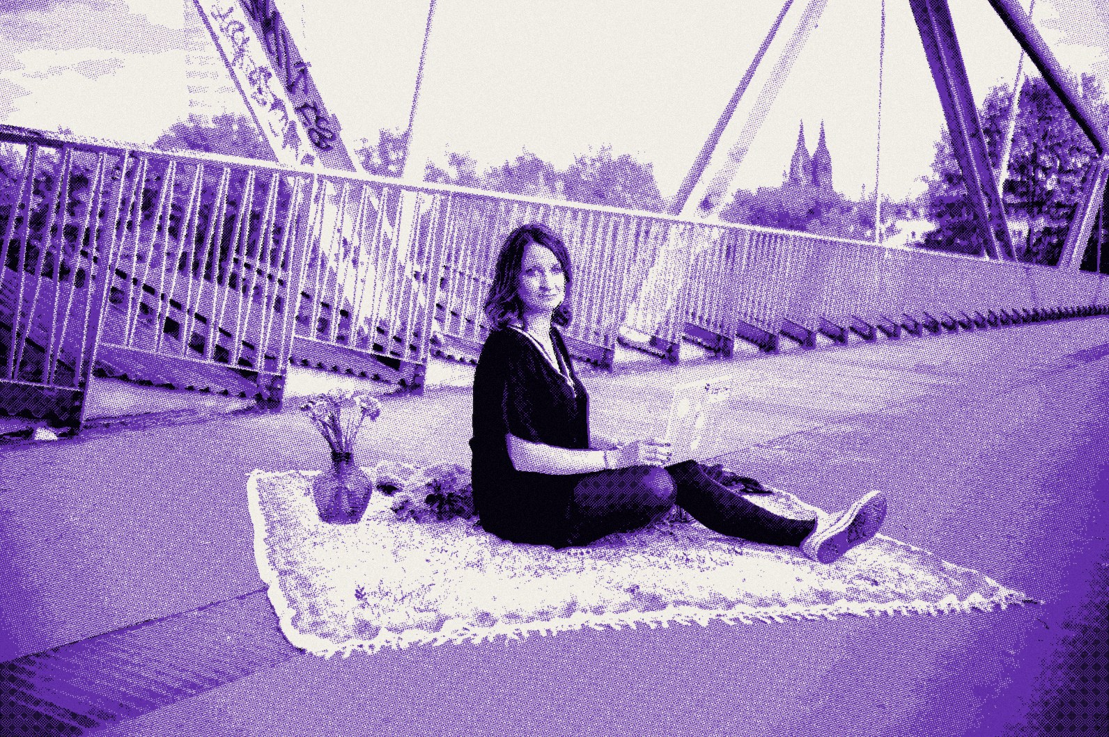 Raphaela Ayriss sitzt seitlich auf einem Teppich auf einer Brücke, hinten der Kölner Dom